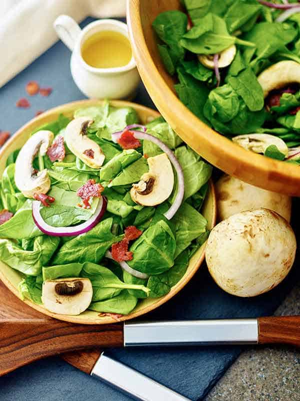 plate of warm bacon spinach salad next to larger bowl above