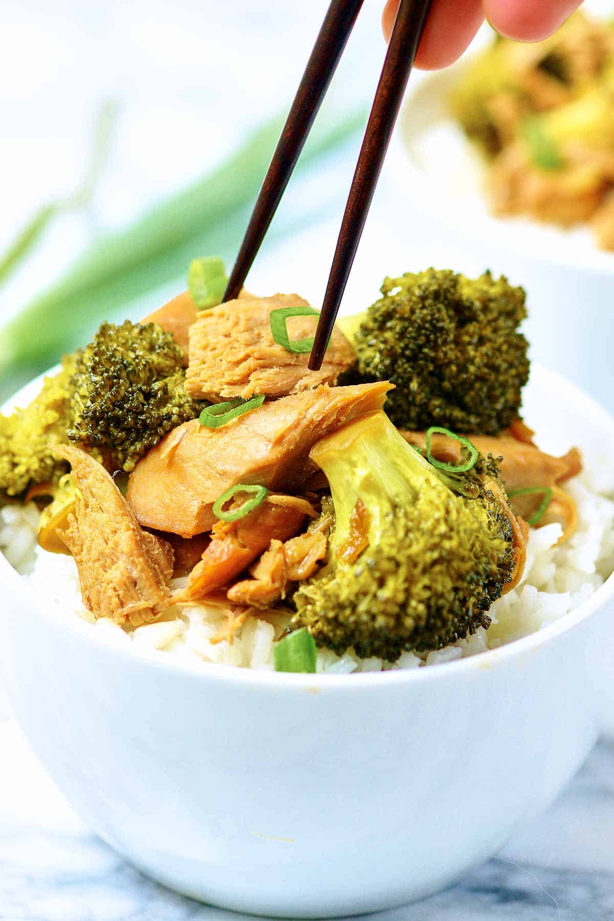 Chicken being taken out of bowl of chicken, broccoli, and rice with chopsticks