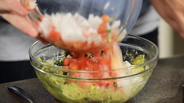 Veggies being poured into bowl of mashed avocado