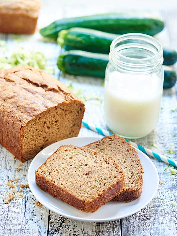 two slices of zucchini bread on plate next to loaf and mason jar of milk