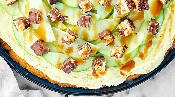 The BEST fall dessert: Snickers Caramel Apple Dessert Pizza. A giant peanut butter cookie topped with a fluffy caramel frosting, apples, and Snickers candy! showmetheyummy.com #snickers #caramel