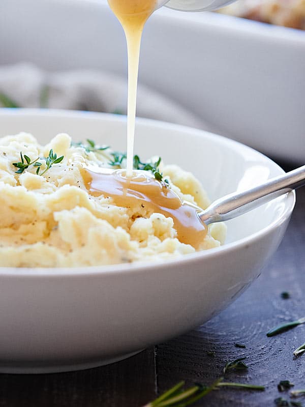 Gravy being drizzled over crockpot mashed potatoes in a bowl with spoon