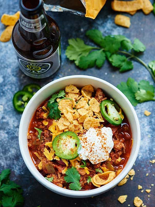 Bowl of chili with jalapeno slices, chips, and sour cream next to beer bottle above