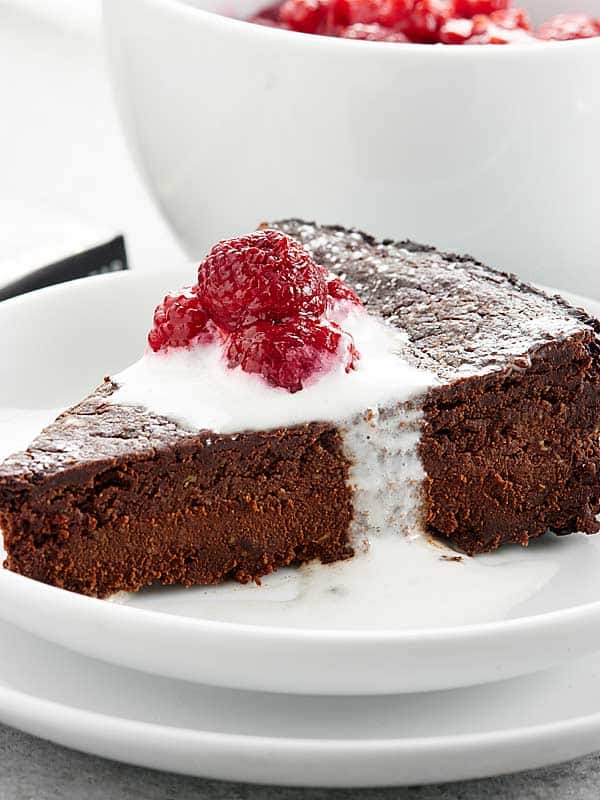 Slice of vegan flourless chocolate cake on plate with icing and raspberries