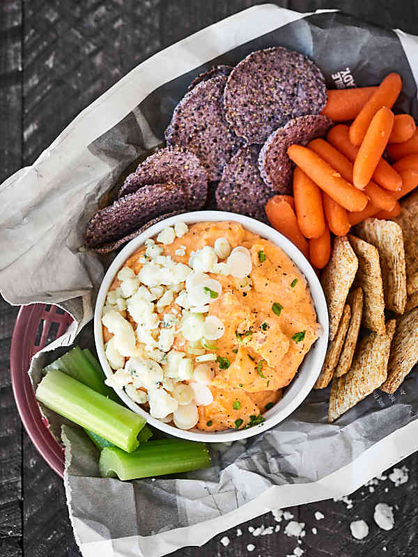 slow cooker cheesy buffalo chicken dip in bowl with basket of veggies, crackers, and chips above