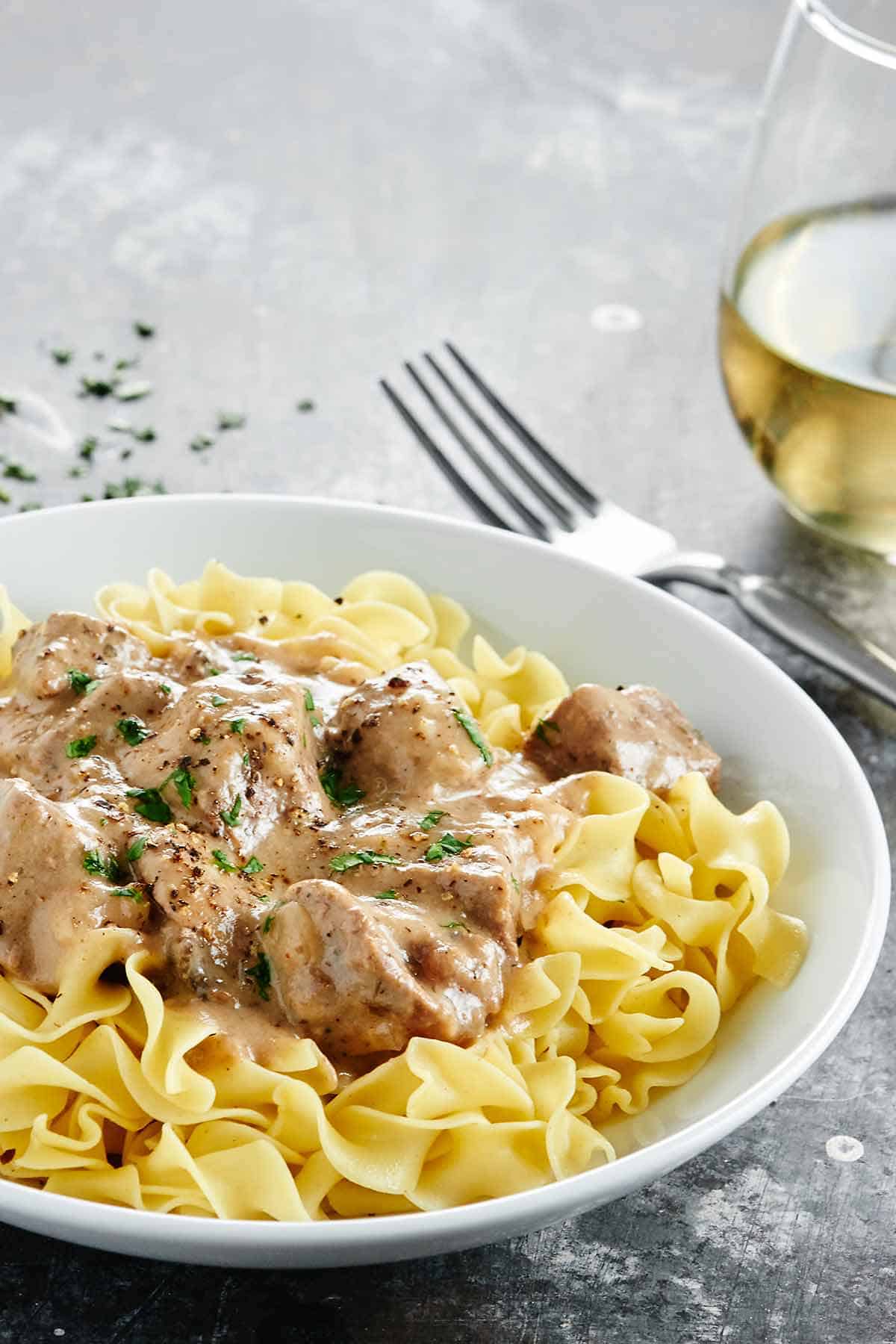 Crock pot beef stroganoff with egg noodles on a plate. Cup of white wine in background