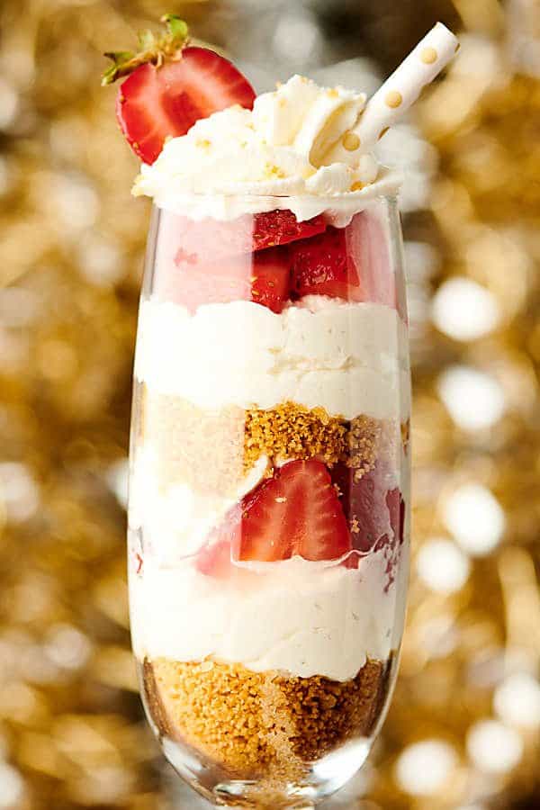 Close-up side shot of no-bake champagne cheesecake in champagne flute with straw and sliced strawberry on top.