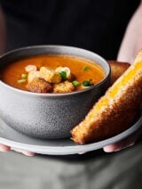 Bowl of roasted tomato soup on a plate with grilled cheese sandwich