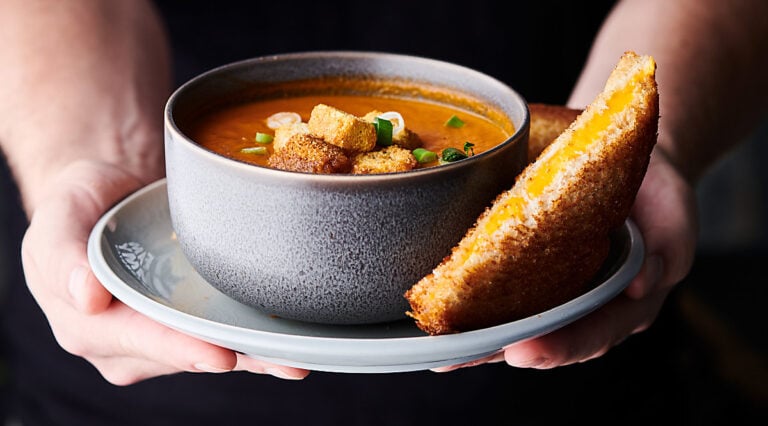 Bowl of roasted tomato soup on a plate with grilled cheese held