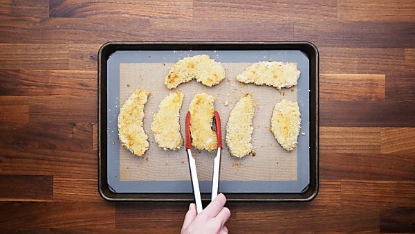 Finished chicken tenders; one being lifted off tray with tongs