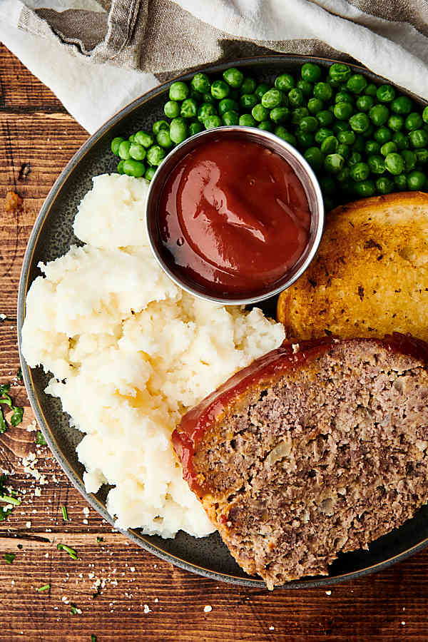 Plate of meatloaf, mashed potatoes, ketchup, peas, and texas toast above