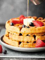 3 waffles stacked with blueberries, strawberries, butter, and maple syrup being drizzled