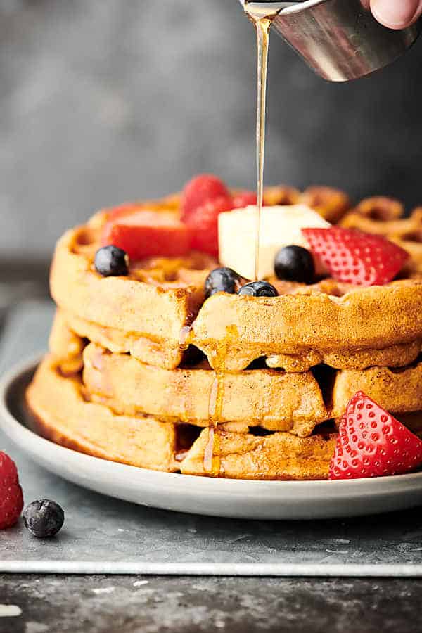 3 waffles stacked with blueberries, strawberries, butter, and maple syrup being drizzled