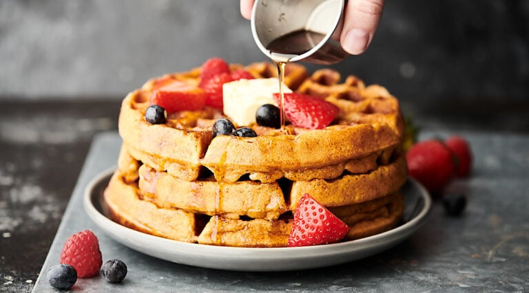 3 waffles stacked, topped with mixed berries, whipped cream, and being drizzled with maple syrup