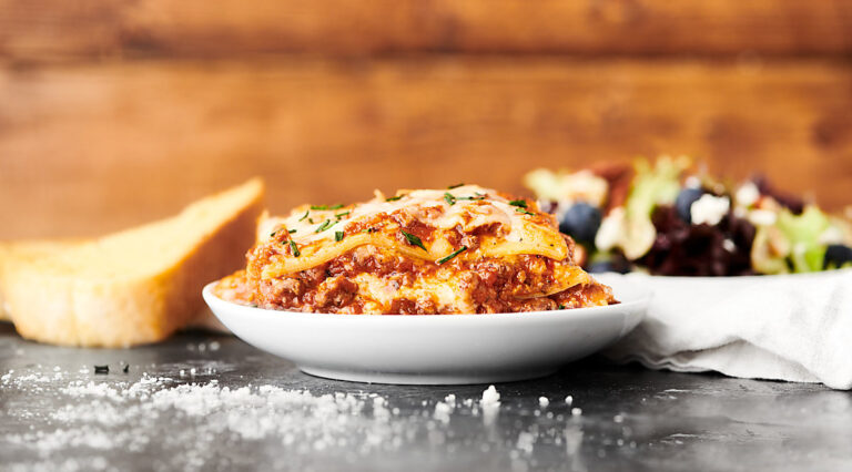 lasagna on a plate, garlic bread and salad in background