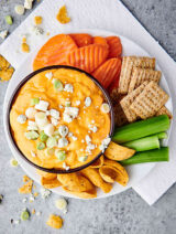 bowl of instant pot buffalo chicken dip on plate with carrots, crackers, celery, and chips above