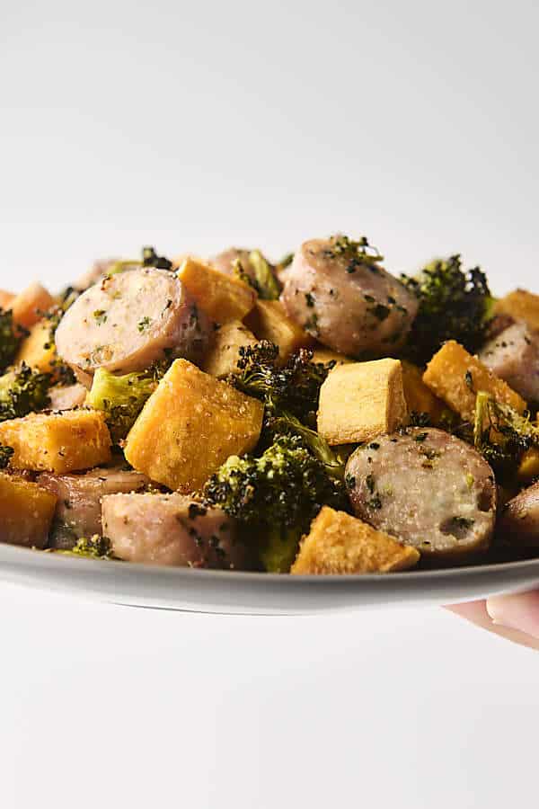 holding a plate of roasted chicken sausage with broccoli and sweet potato 